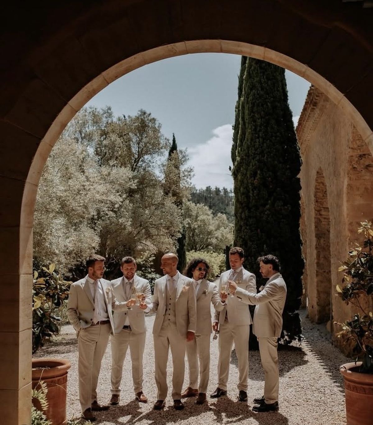 Groomsmen with drinks framed by the stone archway, cypress and terracotta pots beyond