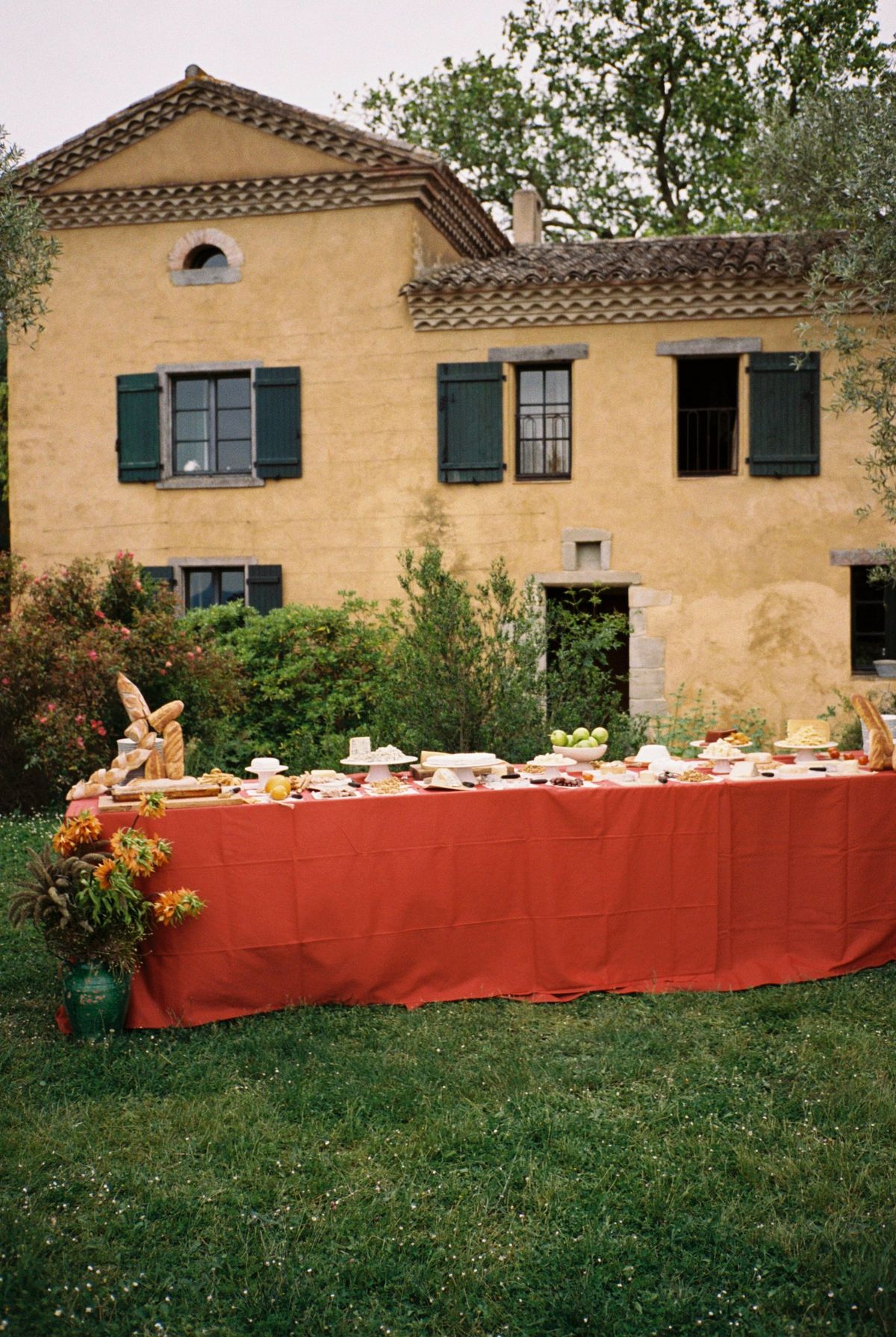 Long buffet table dressed in red linen, laid with bread and cheeses before the ochre farmhouse