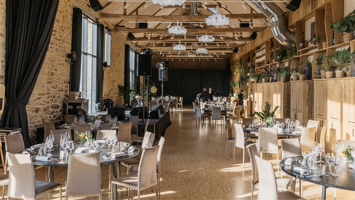 The stone reception hall at Domaine Le Mezo set with round tables and a stage beneath exposed oak beams