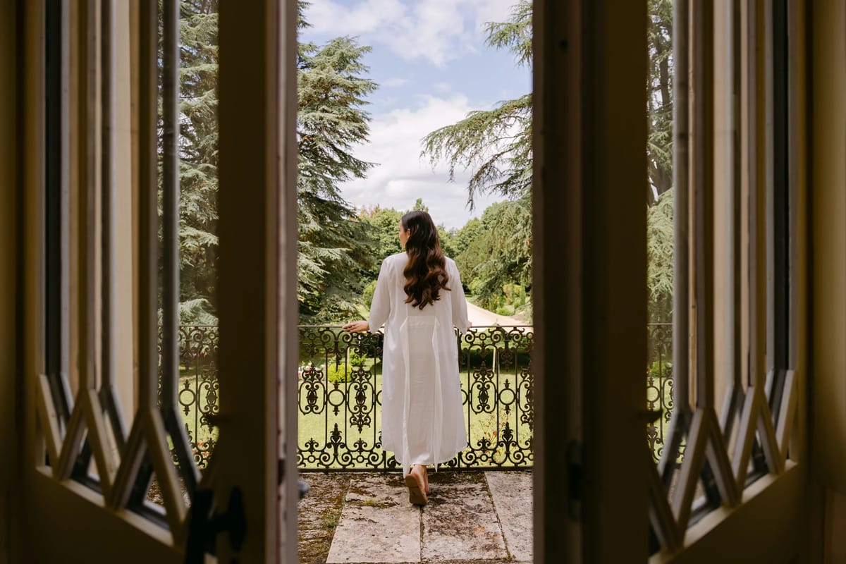 A woman in white standing on a balcony, seen through open shutters, looking out over the garden