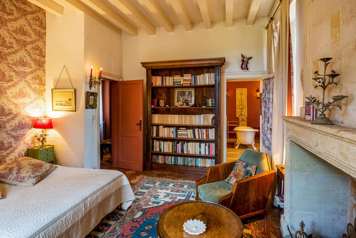 A bedroom with a toile wall, a bookcase, a stone fireplace and a green armchair