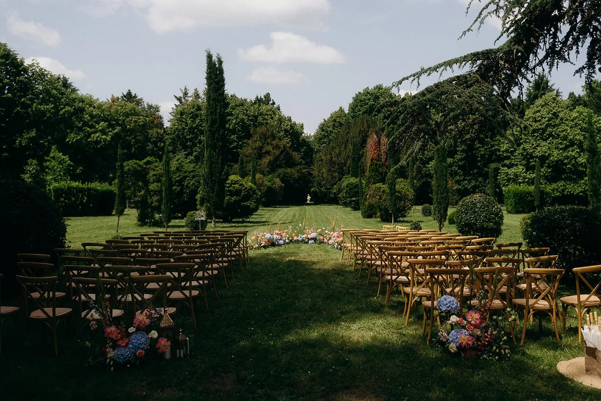 Wooden ceremony chairs set in two blocks on the lawn, with flowers lining the aisle between cypress trees