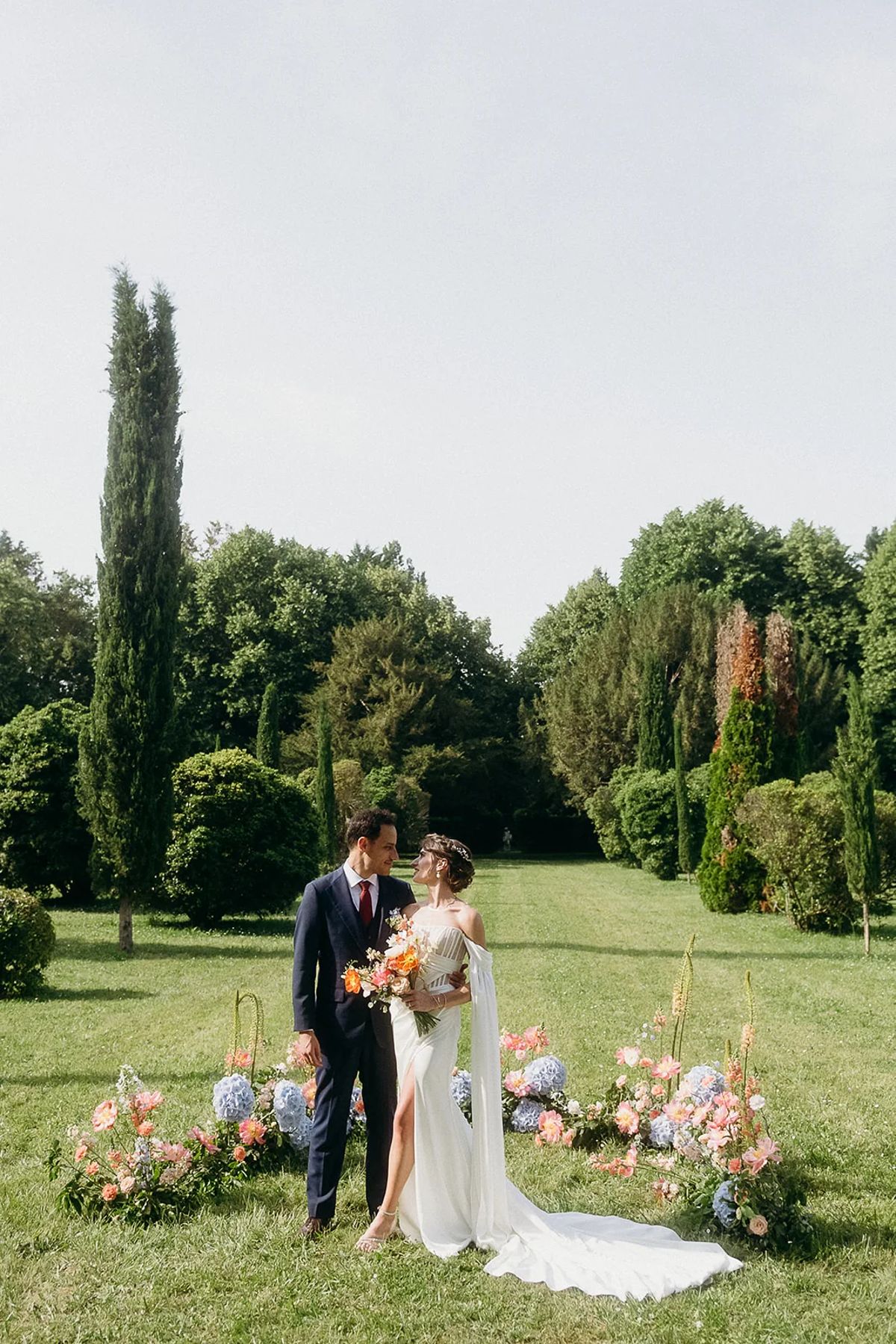 A couple embracing on the lawn in front of low floral arrangements, with cypress and woodland behind