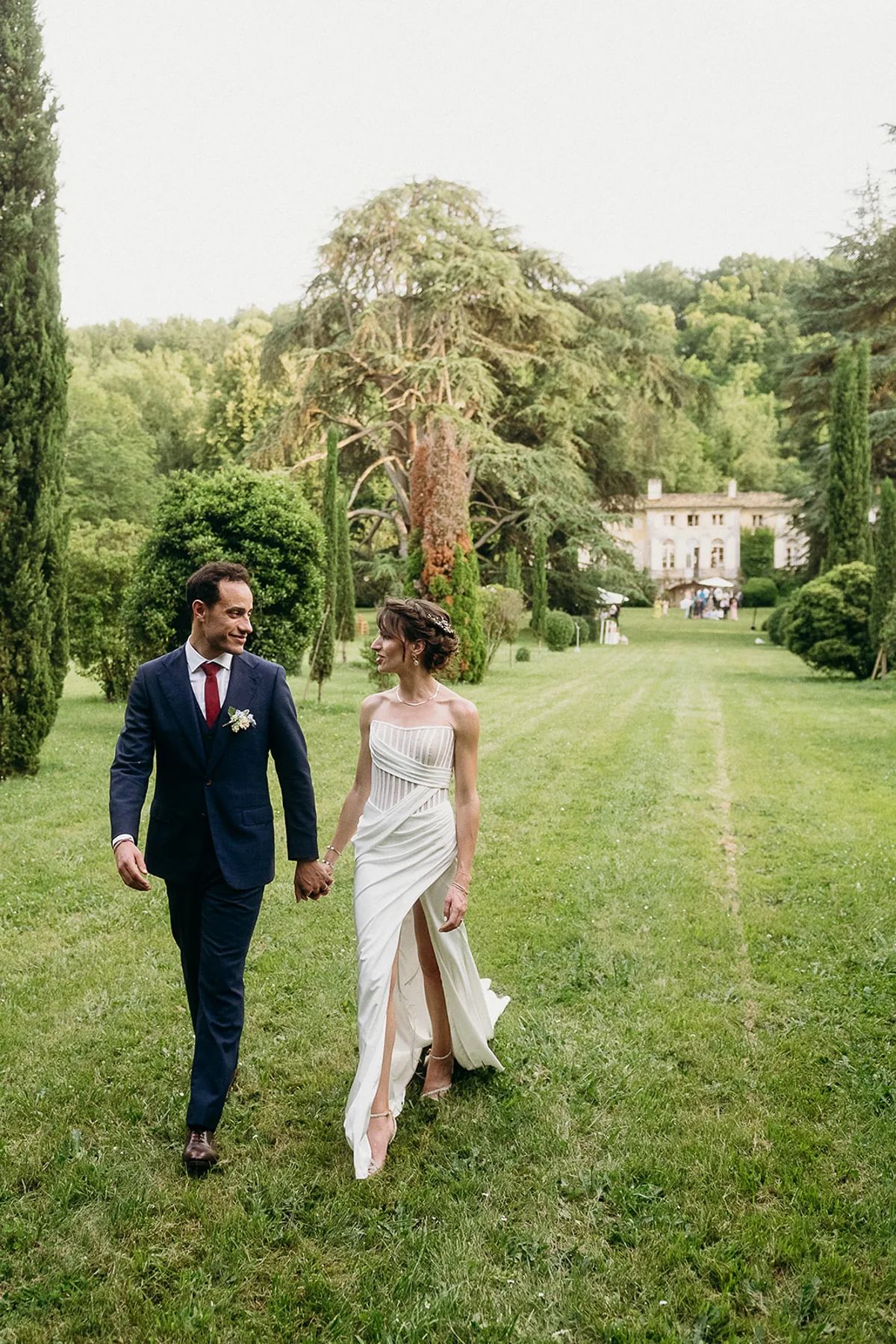 A couple walking hand in hand across a mown lawn with tall cedars and the house in the distance