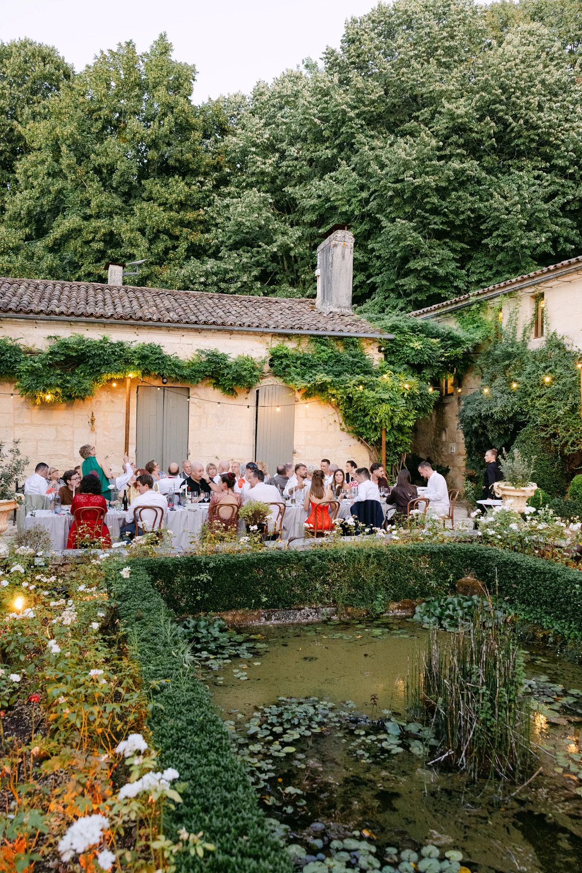 Wedding guests seated at long tables in the courtyard, beyond a hedged lily pond