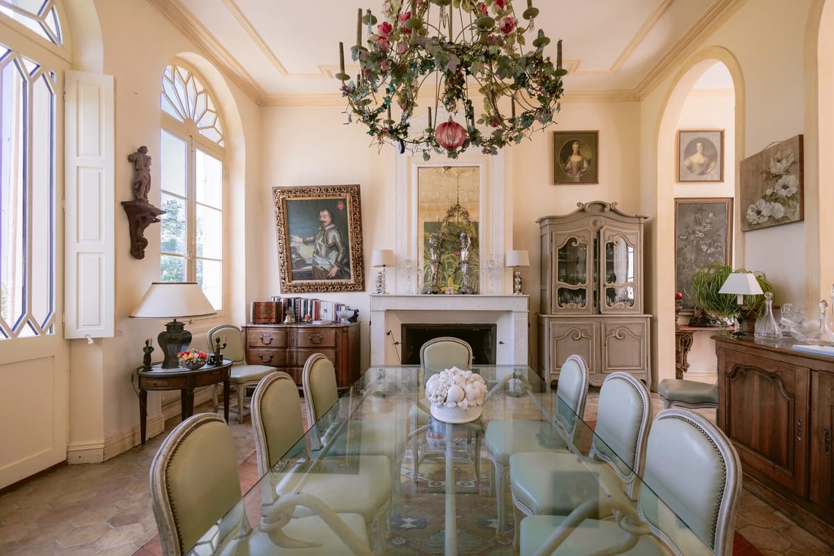 A dining room with a floral chandelier, a glass-topped table and arched windows