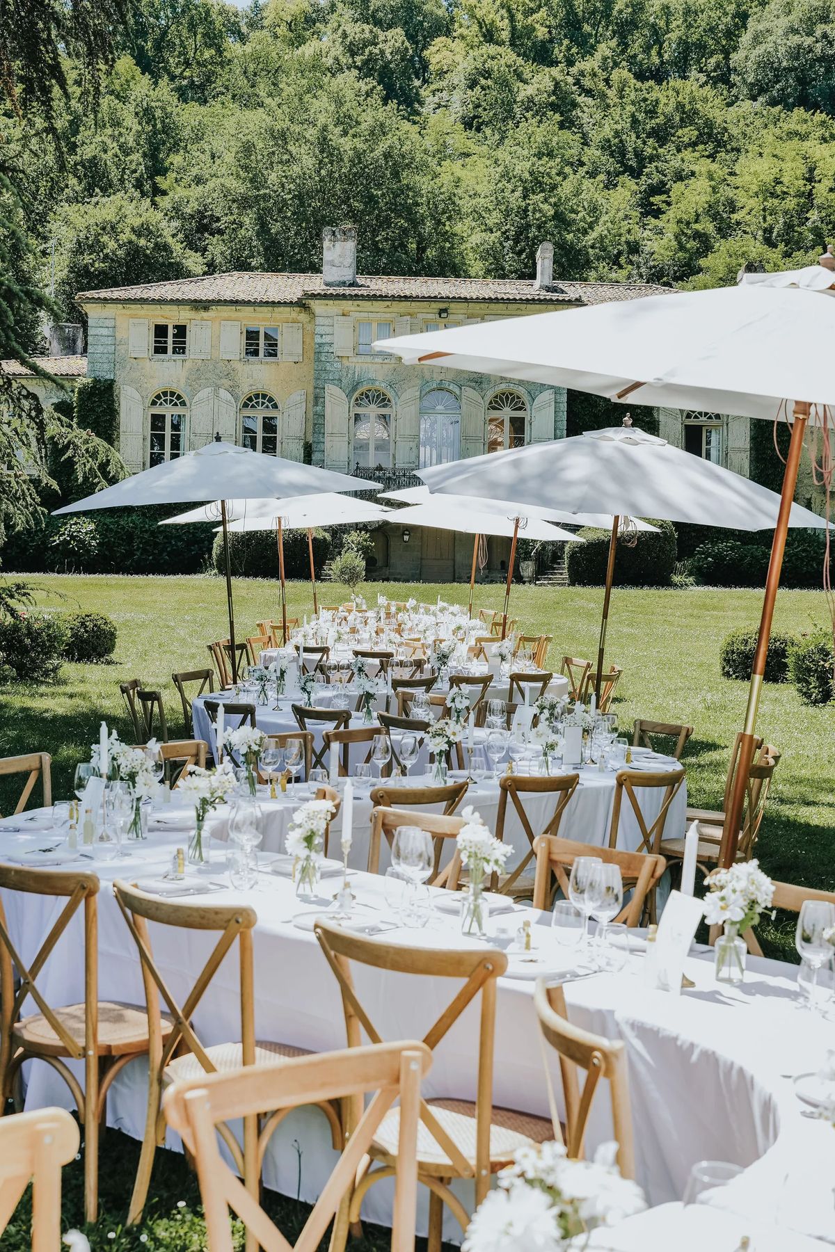 Round tables dressed in white and shaded by parasols on the lawn, with the house behind