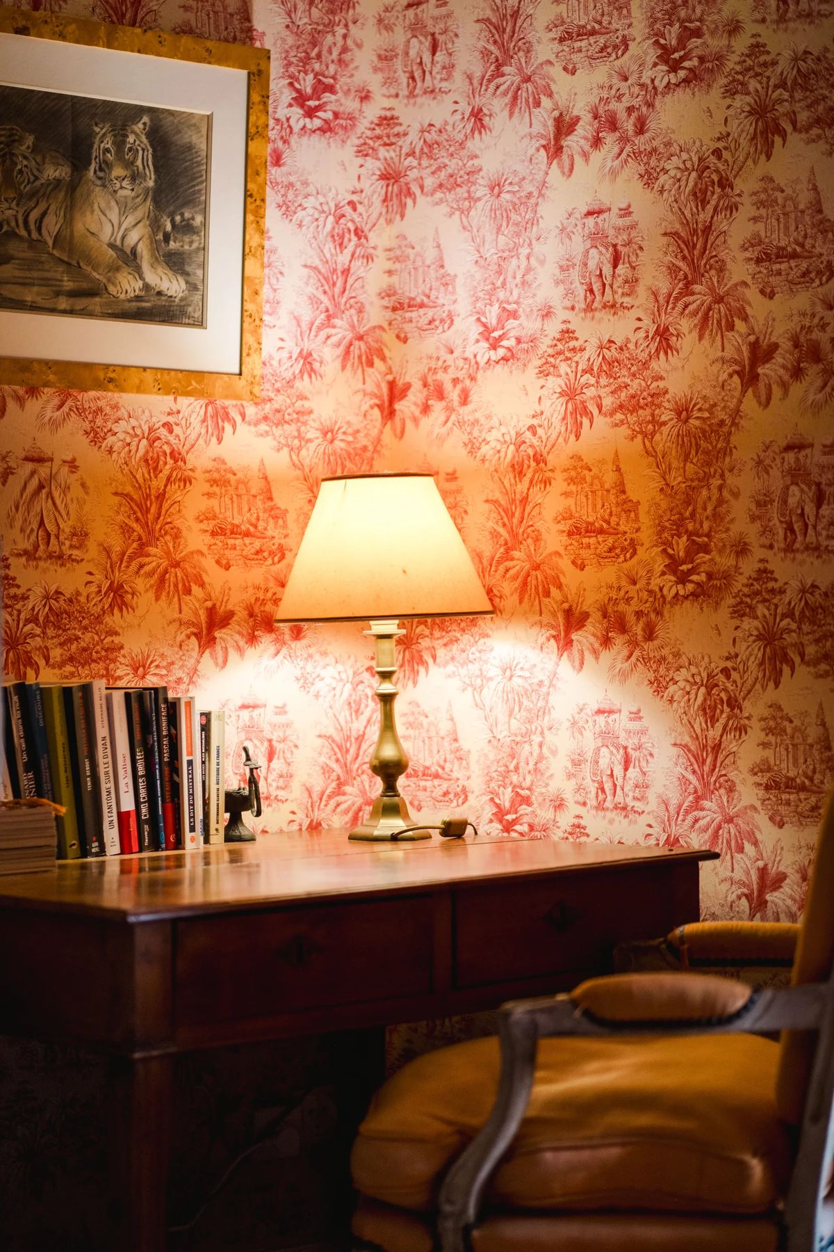 A lamp on a wooden desk against red toile wallpaper, with books alongside