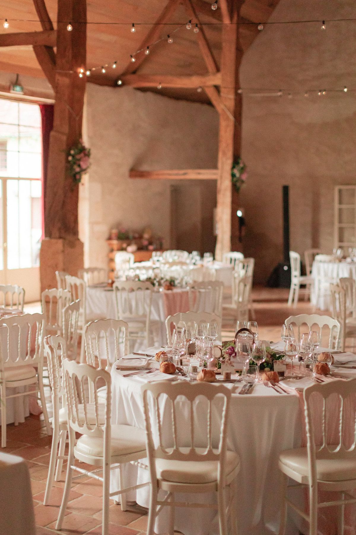 Round tables in white linen with Napoleon chairs and festoon lights under the oak beams of the barn