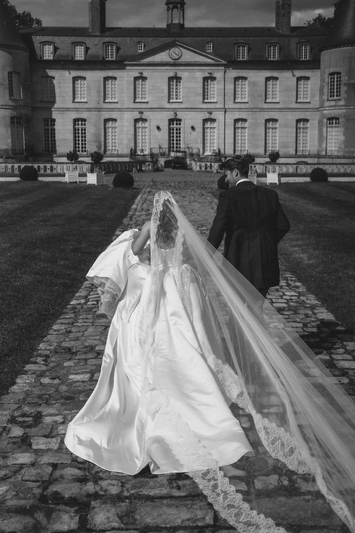 A bride in a long lace-edged veil and a groom walking up the cobbled drive to the château, in black and white
