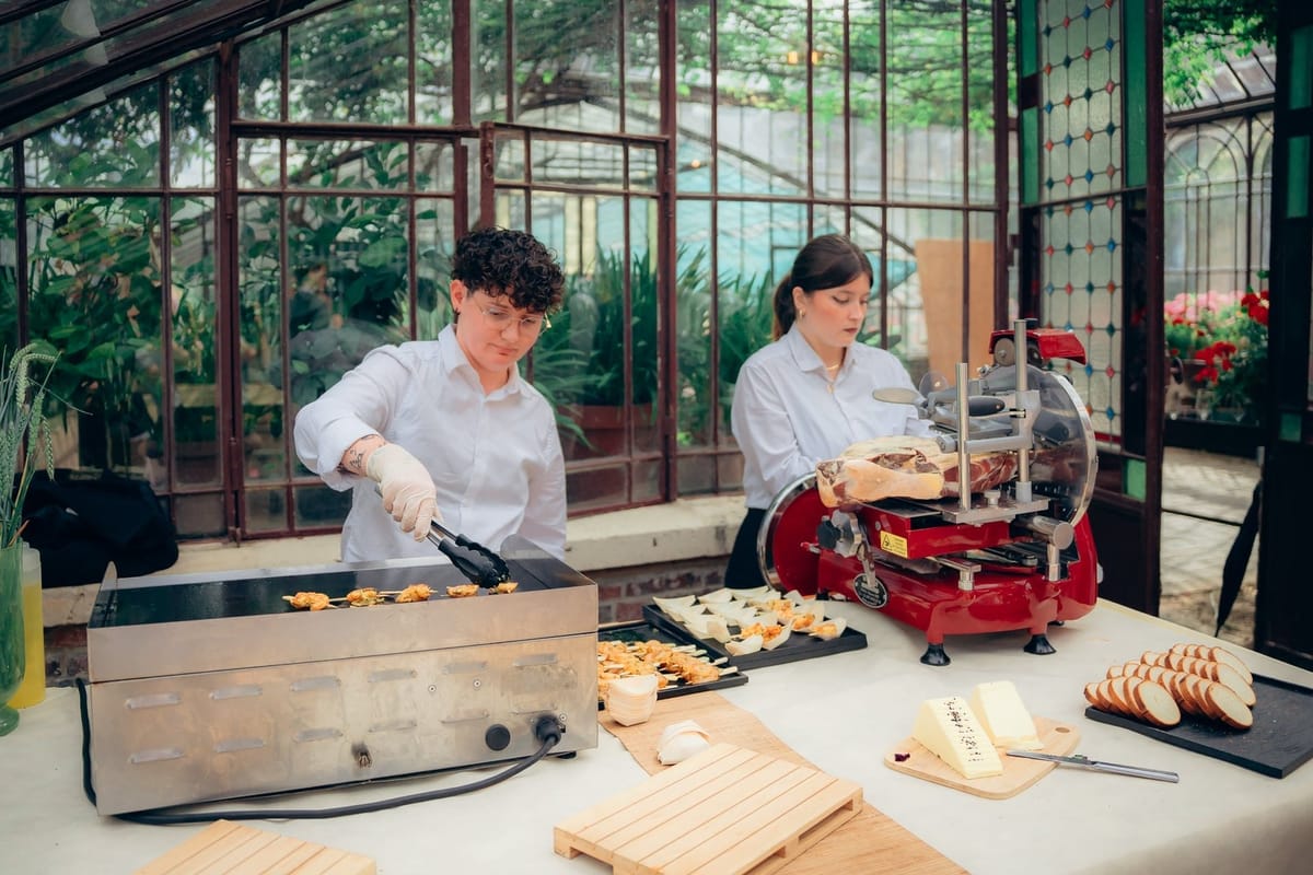 Two catering staff at a food station in the glass greenhouse, one grilling skewers, one at a red ham slicer
