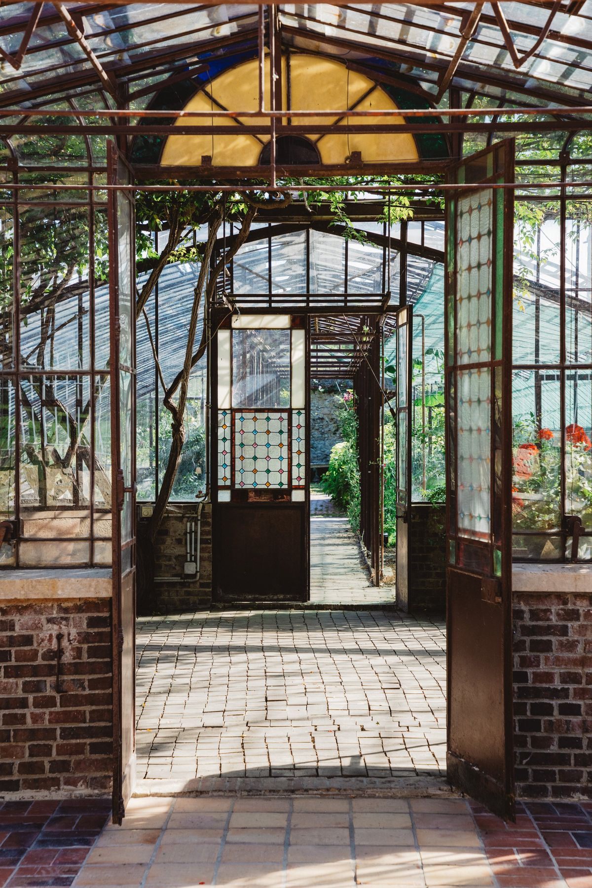 Open greenhouse doors with stained-glass panels leading through to a brick-floored glass corridor