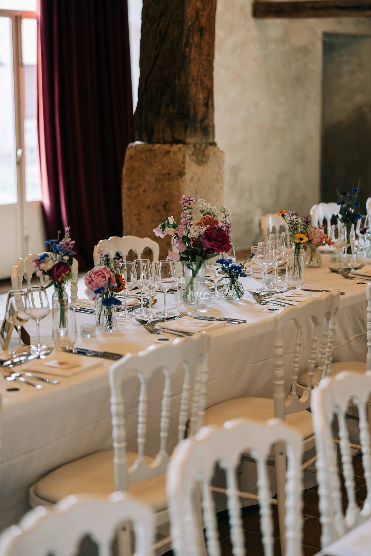 A long table in the barn laid with glassware and small vases of peonies, cornflowers and wildflowers