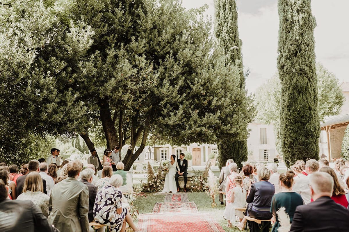 Wedding ceremony at Mas D'Arvieux with couple under olive tree, cypress trees, and vintage rug aisle