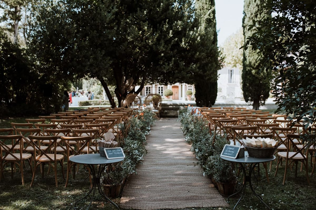 Mas D'Arvieux outdoor ceremony setup with cross-back chairs and olive greenery aisle under cypress trees