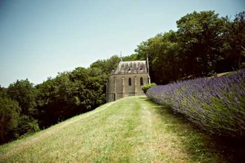 Wide-angle exterior shot of a small historic stone chapel situated on a hillside, with a long row of blooming purple lavender lining a path or wall in the foreground right. No people are visible in the image. The chapel features Gothic-style architecture with pointed rooflines and arched windows. The shot appears to be a venue grounds photograph taken in bright midday natural light. Potential venue feature image.