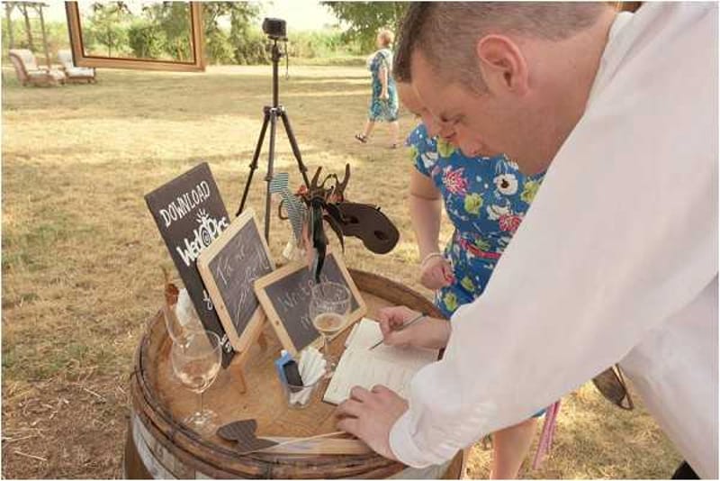A male guest in a white shirt leans over a rustic wooden barrel used as a cocktail-hour guest book station, writing a message in an open notebook. The barrel is styled with small chalkboard signs on easels , one reads 'Download WeddingPics' , along with two wine glasses and a bottle, giving the setup a casual, rustic feel. A camera on a tripod is positioned nearby, likely set up as a photo booth, and a woman in a blue floral dress walks in the background through an outdoor garden or grounds setting. The composition is a medium close-up shot taken in natural daylight.