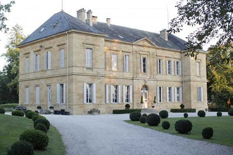 Wide exterior shot of a classic French château, a two-story limestone building with a slate mansard roof, white shuttered windows, and a central arched entrance door with interior lighting visible. The grounds feature a gravel driveway flanked by neatly clipped spherical boxwood topiaries and manicured lawn. No people are present in the image. Potential venue feature image.