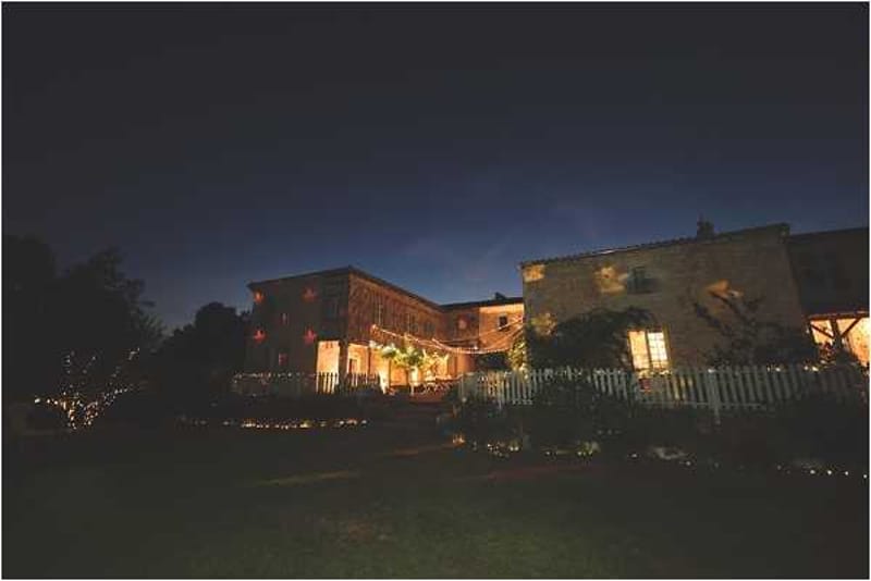 A wide-angle nighttime exterior shot of a rustic stone farmhouse or barn venue illuminated by warm amber interior lighting and string lights draped across the facade and surrounding trees. The venue features multiple interconnected stone buildings with lit windows, a white picket fence, and what appears to be an outdoor terrace or covered area strung with festoon lights. Small ground-level lights line a pathway in the foreground, creating a warm glow against the deep blue twilight sky. No people are visible in the frame. Potential venue feature image.