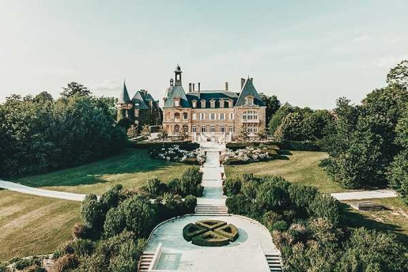 Aerial wide shot of a French château with classic 19th-century architecture, featuring brick facades, slate-roofed turrets, and dormer windows. The formal grounds include a symmetrical garden approach with a circular parterre in the foreground, a central pathway leading up to the building, manicured hedgerows, and white flower beds flanking the main steps. No people are visible in the image. Potential venue feature image.