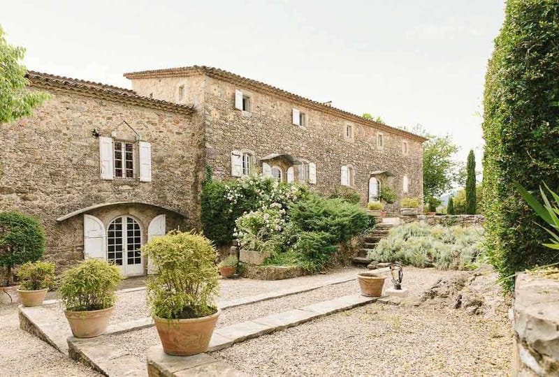 Wide exterior shot of a large Provençal stone farmhouse or mas, featuring traditional terracotta roof tiles, white-shuttered windows, and an arched double French door entrance. The foreground shows a gravel courtyard with terracotta pots planted with topiary shrubs and ornamental plants, along with stone steps leading up to a terraced garden area. White flowering shrubs are visible growing against the facade, and cypress trees frame the right side of the building. No people are present in the image. Potential venue feature image.