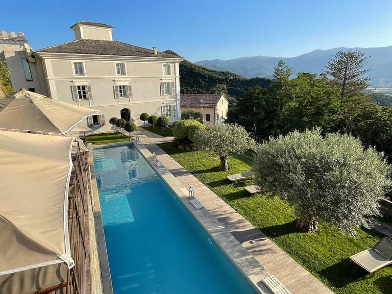 An aerial-angle exterior shot of a large white manor house with grey shutters and a slate roof, viewed from an elevated terrace position. In the foreground is a long rectangular lap pool with a wooden deck surround, flanked by sun loungers and glass lantern decorations. The grounds include a manicured lawn with topiary ball-shaped trees lining a pathway toward the main building, as well as mature olive trees. A beige shade sail or awning structure is visible on the left edge of the frame. The setting is a hillside property with mountain views in the background. No people are present in the image. Potential venue feature image.