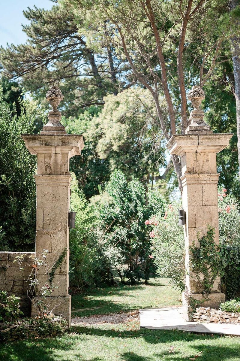 An outdoor venue shot featuring a pair of tall, weathered limestone gate pillars topped with carved stone urn finials, forming an open entrance to a garden. A stone pathway leads through the gateway, flanked by low stone walls and lush garden plantings including shrubs and what appear to be pink-flowering oleanders to the right. No people are present in the frame. The wide shot is taken in bright midday light with a classic, formal architectural style typical of a French estate or chateau property. Potential venue feature image.