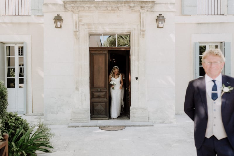 The bride is stepping out through a large wooden double door of a French chateau or manor house, captured in a wide shot that includes the full facade of the building. She is wearing a fitted white lace gown with a V-neckline and a long veil, and is holding a bouquet of white flowers, likely white roses or peonies. In the foreground to the right, an older man in a navy suit with a light grey waistcoat and navy tie — likely the father of the bride — stands with his back partially to the camera, out of focus. The building exterior features pale stone, blue-grey shutters, and two wall-mounted lanterns flanking the ornate doorway. Potential venue feature image.