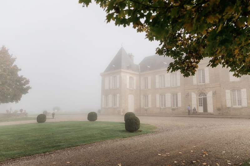 Wide exterior shot of a French chateau photographed on a heavily foggy day, with the stone building partially obscured by mist. The chateau features classic French architecture with a steep slate-roofed central tower, white-shuttered windows, and an arched entrance doorway. The formal grounds in the foreground include a gravel driveway, a manicured lawn, and several rounded clipped boxwood topiaries. Two people are visible standing near the entrance to the right of the building. Tree branches with autumn-toned foliage frame the upper right corner of the composition. Potential venue feature image.