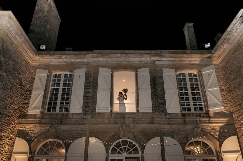 A bride in a fitted white lace-back gown stands in an illuminated upper-floor window of a French chateau at night, holding her bouquet of greenery raised above her head. The wide-angle shot is taken from ground level looking up at the chateau facade, which is warmly lit by exterior uplighting and features white shuttered windows and arched ground-floor openings strung with fairy lights. The contrast between the glowing interior light silhouetting the bride and the deep black night sky creates a dramatic compositional effect. Potential venue feature image.