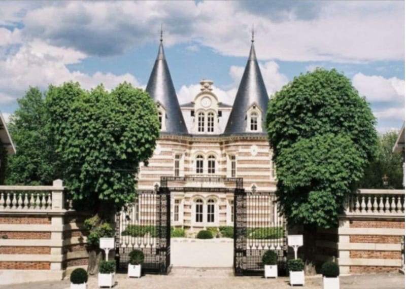 A wide exterior shot of a French château featuring two pointed slate-grey turrets, ornate red brick and cream stone striped façade, and arched windows with a central clock detail. The entrance is framed by wrought iron gates flanked by white planters with clipped boxwood topiaries, and a low brick balustrade runs along the front perimeter. No people are visible; the image focuses entirely on the architecture and grounds approach. Potential venue feature image.