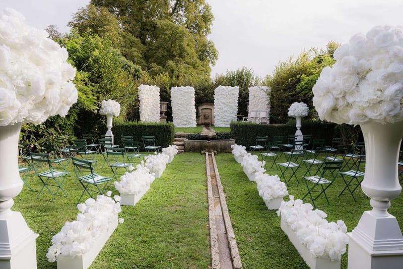An outdoor wedding ceremony setup photographed before guests arrive, shot from a wide angle at the entrance of the aisle. The ceremony space is set within a formal garden featuring a central stone water channel running down the middle of the lawn, flanked by two rows of dark green metal folding chairs arranged in a curved layout. The aisle is lined with white floral arrangements — large bunches of white roses and hydrangeas — displayed in white rectangular planters. At the entrance, two oversized white urn pedestals hold large spherical arrangements of white roses and hydrangeas. The altar backdrop consists of three white flower walls composed of densely packed white roses, framing a classical stone fountain with a sculpted face mask, set against a neatly trimmed tall hedge. Two additional white floral topiary arrangements on white pedestal urns flank the altar area. The overall decor palette is entirely white against deep green, creating a classic formal style. Potential venue feature image.