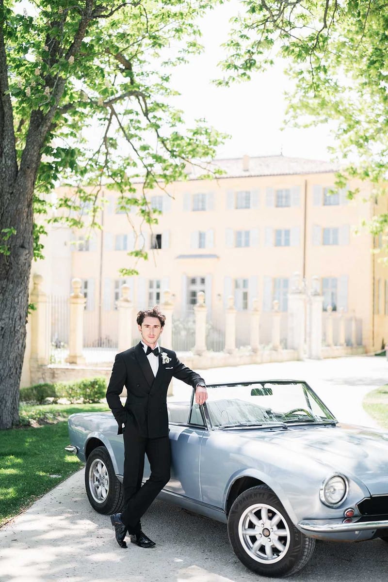 A groom portrait taken outdoors on the driveway of a French chateau, with the pale yellow facade of the manor house softly out of focus in the background. The groom is dressed in a black double-breasted tuxedo with a white dress shirt, black bow tie, white pocket square, and a small white floral boutonniere, leaning casually against a vintage powder blue convertible sports car. The shot is a full-length portrait with a classic, formal styling aesthetic. Potential venue feature image.