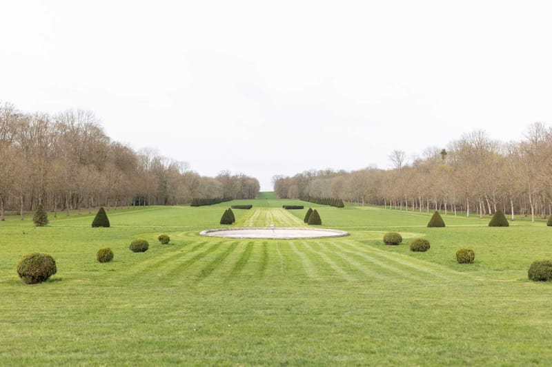 Wide shot of formal French-style estate grounds featuring a long, symmetrical allée extending toward the horizon, flanked by bare-branched trees on both sides. In the foreground, a circular stone fountain basin sits at the center of a striped mown lawn, surrounded by cone-shaped and rounded topiary shrubs arranged in a formal pattern. No people are visible in the image. Potential venue feature image.
