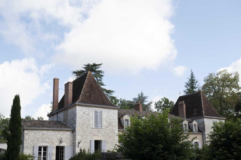A wide exterior shot of a traditional French country manor house, featuring cream-colored stone facades, dark brown tiled steeply pitched rooftops with dormer windows, white-shuttered windows, and brick chimneys. The building spans multiple connected wings with conical tower rooflines typical of French regional architecture. No people or wedding decor are visible in this image. Potential venue feature image.