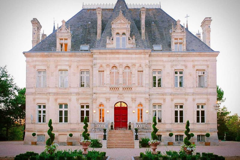 A wide exterior shot of a French château at dusk, showing the full symmetrical facade of a three-story limestone building with a slate mansard roof, ornate stone carvings, and dormer windows. The entrance features a bold red double door framed by an arched transom window, flanked by warm wall-mounted lanterns that cast a golden glow, with a broad stone staircase leading up to the entrance. Formally trimmed spiral and conical topiary shrubs are arranged symmetrically along the front facade, and terracotta urns planted with white flowering arrangements flank the base of the steps. No people are visible in the frame. Potential venue feature image.