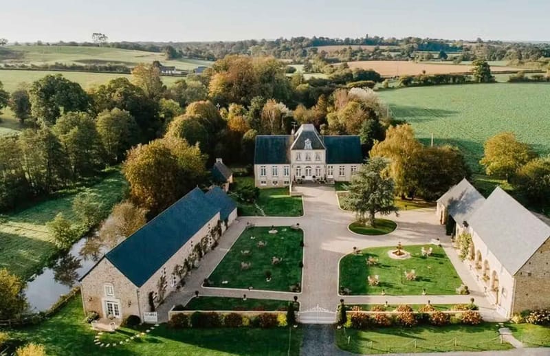 Aerial drone shot of a French chateau estate showing the full property layout, including a central manor house with a slate mansard roof and cream stone facade, flanked by two large outbuildings — one with a blue slate roof on the left and a stone barn with arched arcade detailing on the right. The grounds feature two formal parterre gardens with trimmed lawns, a circular driveway with a central planted roundabout, a white iron entrance gate, topiary accents, and flower borders in warm orange and red tones suggesting an autumn setting. A small ornamental pond is visible to the left of the main outbuilding. The estate is surrounded by open agricultural fields and mature trees with autumn foliage in gold and amber. No people are visible in the frame. Potential venue feature image.