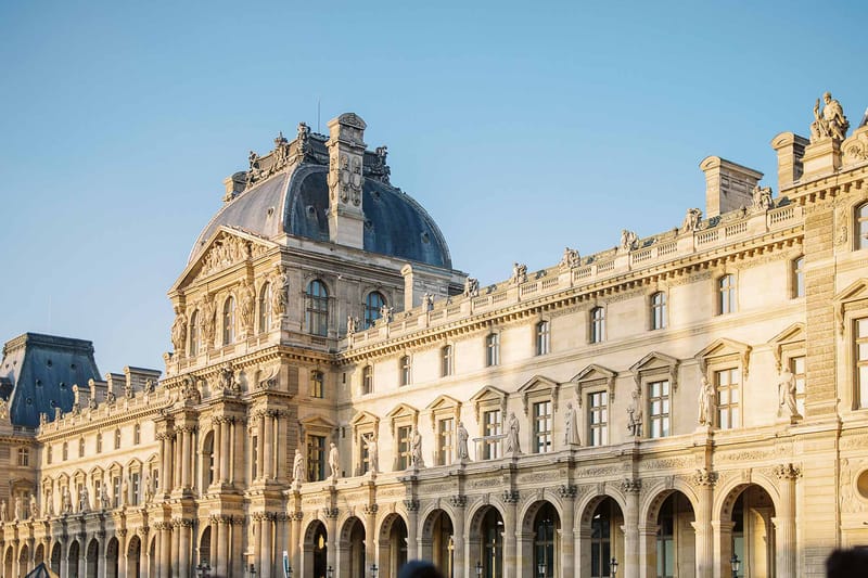 A wide-angle exterior shot of the Louvre Palace courtyard facade in Paris, showing the classic French Renaissance architecture with its cream-colored stone, ornate sculptural detailing, arched ground-floor colonnades, and a dark slate-domed pavilion rising at the wing junction. The building features multiple tiers of tall arched windows, relief sculptures, and full-figure statues positioned between bays across two upper floors. No people, couple, or wedding activity are visible in this image — it functions purely as an architectural location shot. Potential venue feature image.
