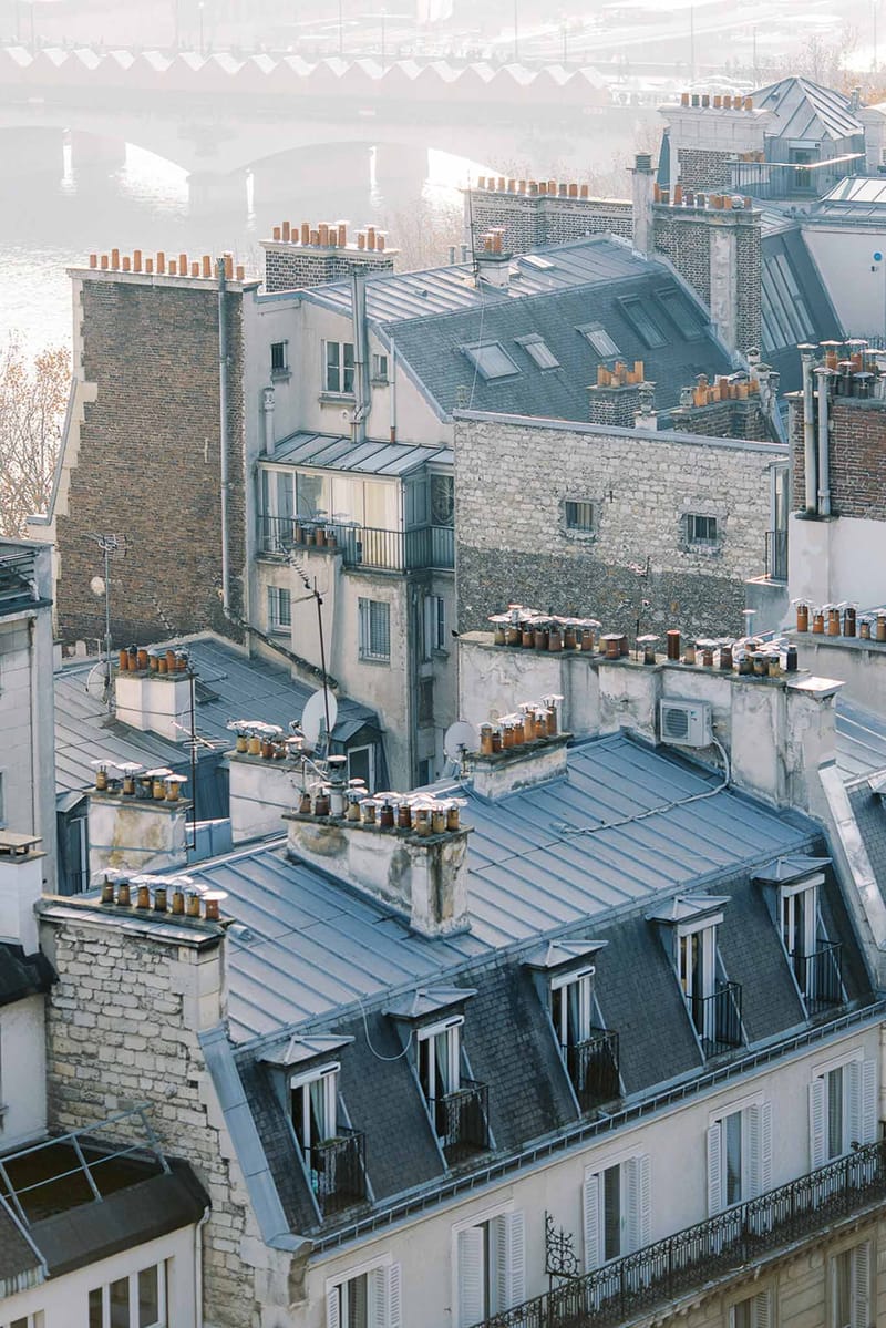 This image contains no wedding-related content. It is an aerial/elevated wide shot of Parisian rooftops featuring classic zinc-grey mansard roofs, terracotta chimney pots, dormer windows, and Haussmann-style building facades in cream and stone. A river — likely the Seine — and a bridge are visible in the misty background. There are no people, couples, wedding attire, florals, or event decor present in the frame.