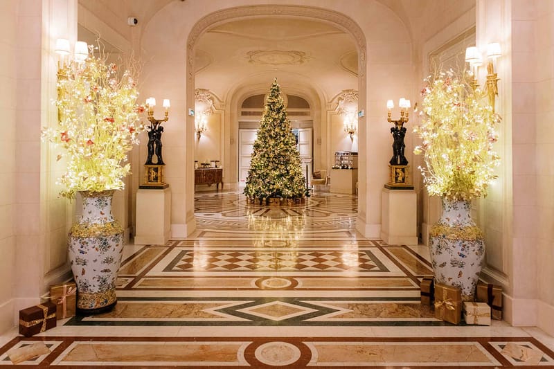 Wide shot of an indoor grand hotel or palace lobby decorated for the winter holiday season, with no people visible. The space features a polished geometric marble floor in cream, green, and terracotta tones, arched doorways with ornate molded ceilings, and warm wall sconces mounted on black and gold figural candelabra pedestals. Two large white porcelain vases with floral and butterfly motifs flank the corridor, each filled with tall illuminated branches in yellow-green and coral tones. A large Christmas tree decorated with warm white fairy lights and gold ornaments stands centered in the background, surrounded by wrapped gifts in gold and brown. The overall decor palette is cream, gold, and warm white, with a classic French palatial interior style. Potential venue feature image.