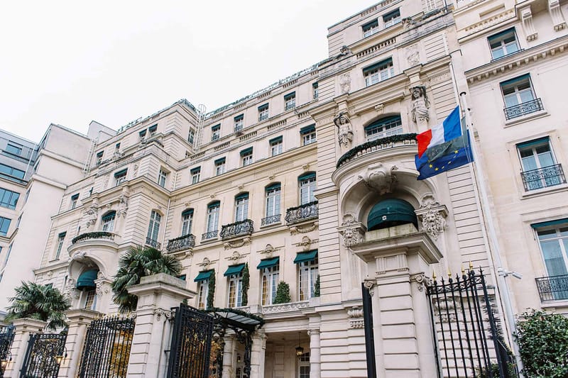Wide exterior shot of a grand Haussmann-style Parisian building facade, photographed from street level looking upward. The cream limestone building features multiple stories with ornate carved stone detailing, sculptural figures, wrought iron balconies, and arched entryways with dark teal awnings. The French tricolor flag and European Union flag are mounted on the right side of the facade above the main arched entrance. Black wrought iron gates and fencing line the front, with palm trees visible in the courtyard to the left. No people or wedding-specific elements are visible in this image. Potential venue feature image.