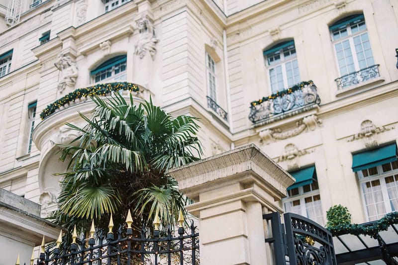 Exterior architectural shot of a grand Haussmann-style Parisian building featuring cream stone facades, ornate sculptural detailing, teal-painted window frames and awnings, and wrought iron balconies with decorative ironwork. The building's balconies and entrance gate are dressed with dark green garland and warm gold fairy lights, suggesting a winter or festive-season event. A large palm tree rises in front of the black and gold-tipped wrought iron entrance gate, with a glazed canopy entrance visible to the right. The composition is an upward-angle wide shot focused on the building's facade and entrance. Potential venue feature image.