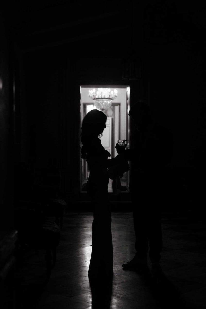 This black-and-white image shows two people silhouetted in a dark interior hallway or anteroom, one holding what appears to be a crystal glass. The figures are backlit by a brightly lit doorway behind them, through which a large crystal chandelier is visible, suggesting a formal chateau or grand mansion interior. The high contrast between the deep black foreground and the bright, warmly lit room beyond creates a dramatic tonal range. The wide shot captures the tiled floor reflecting a small amount of light, and a third person is partially visible crouching in the lower left corner.