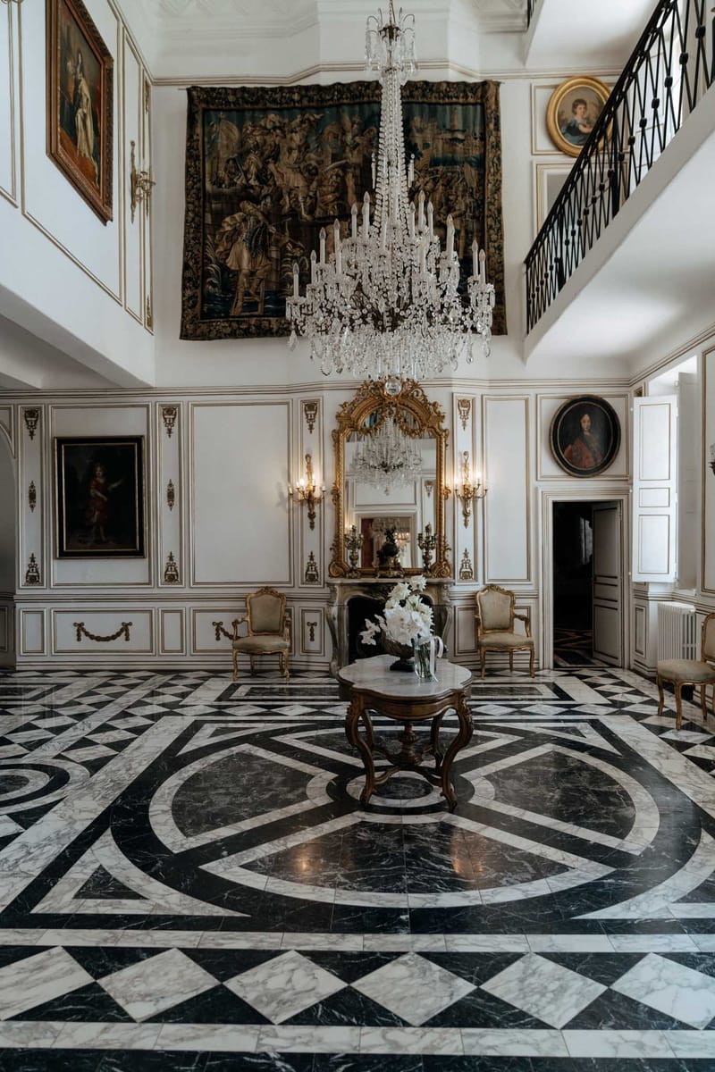 A wide interior shot of a grand chateau entrance hall or salon, with no people present. The double-height room features white paneled walls with gold gilt detailing, a large multi-tiered crystal chandelier hanging from the ceiling, and a dramatic Flemish-style tapestry mounted on the upper wall. A black and white geometric marble floor with a circular medallion pattern dominates the foreground. At center stands a carved walnut occasional table holding a vase of white amaryllis or lily blooms. A marble fireplace with an ornate gold-framed mirror and flanking gold wall sconces sits against the back wall, with two gold-framed Louis XV-style armchairs positioned on either side. Multiple oil portraits in gilded frames hang throughout the room, and a wrought-iron mezzanine balcony is visible on the upper right. The overall style is classic 18th-century French interior. Potential venue feature image.
