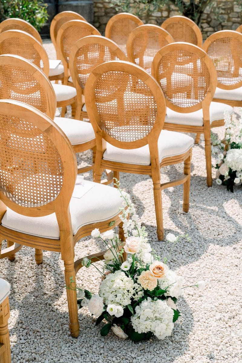 An outdoor ceremony setup shot showing rows of natural wood Louis XVI-style chairs with cane medallion backs and ivory cushioned seats, arranged on a gravel surface. Floral arrangements placed along the aisle feature white hydrangeas, white lisianthus, blush and peach roses, and green foliage. The decor palette is white, blush, and peach with warm natural wood tones, fitting a classic French garden ceremony aesthetic. The composition is a medium angled shot focused on chair and aisle details with no people present.