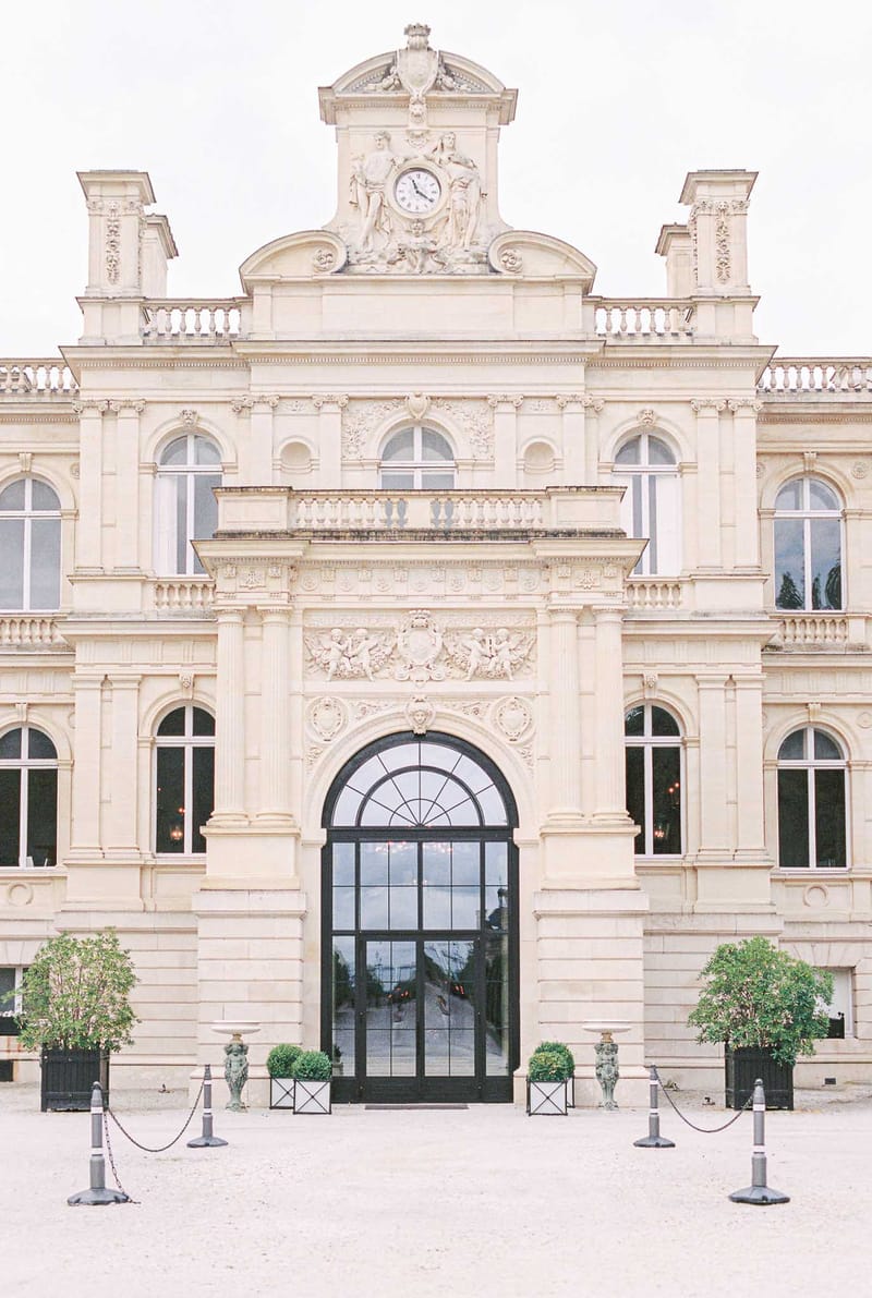 Grand classical chateau facade with bas-relief carvings, pediment clock, gravel forecourt, and conical topiaries