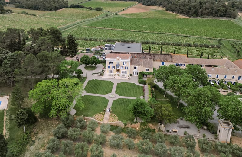 An aerial wide shot of a French domaine or château-style wedding venue, showing a yellow-painted manor house with blue shutters and terracotta roof tiles as the central building, flanked by long stone outbuildings with matching blue shutters. The formal garden in front of the manor features a cross-shaped path layout with four lawn panels and a central stone fountain. Two small figures, appearing to be the couple — one in white — are visible at the base of the garden steps, giving scale to the property. A rectangular swimming pool is visible to the lower left of the frame. The estate is surrounded by extensive vineyards stretching across the upper portion of the image, with rows of vines clearly visible, along with cypress trees and mature deciduous trees framing the property. Potential venue feature image.