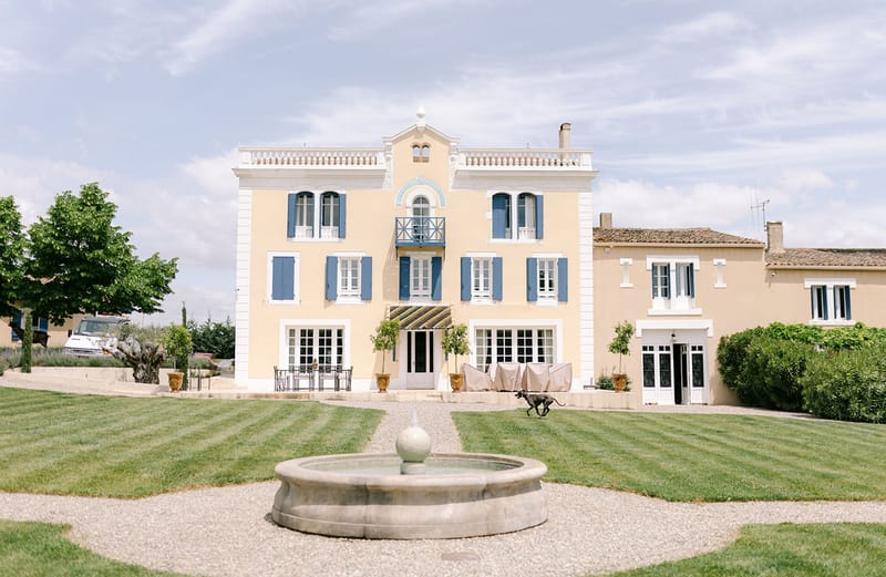 A wide exterior shot of a three-story French manor house with a pale yellow facade, white trim, and blue shutters throughout. The entrance features a colorful geometric-patterned canopy in blue, yellow, and green above the central French doors, flanked by potted topiary trees in terracotta urns. The foreground includes a circular stone fountain with a sphere finial set within a gravel path, surrounded by neatly striped mowed lawn. A dark-colored dog runs across the gravel path in front of the entrance. Outdoor furniture including a metal dining table and chairs is visible to the left of the entrance, with covered furniture to the right. An adjacent lower building in matching yellow render extends to the right. Potential venue feature image.