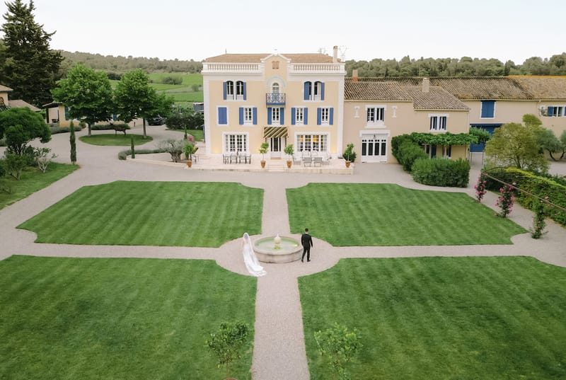 Aerial view of bride and groom walking toward a yellow Provencal manor with formal garden and stone fountain