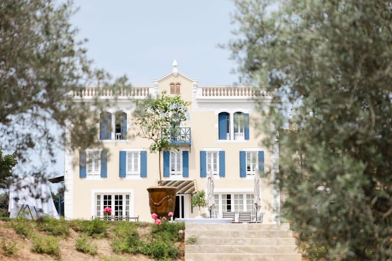 Three-storey Provencal manor with yellow facade, blue shutters, and mosaic-tiled entrance canopy framed by olive trees